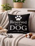 Black pillow with 'Reserved for the DOG' text on a couch with pet toys and a plant in the background.