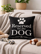 Black pillow with 'Reserved for the DOG' text on a couch with pet toys and a plant in the background.