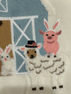 Easter cushion with ambroidered design of a pig, sheep, and rabbit near a barn on fabric for home decor.