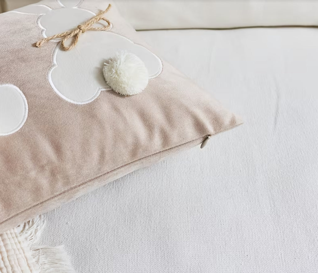 Beige pillow with white cloud designs and a pom-pom on a white surface