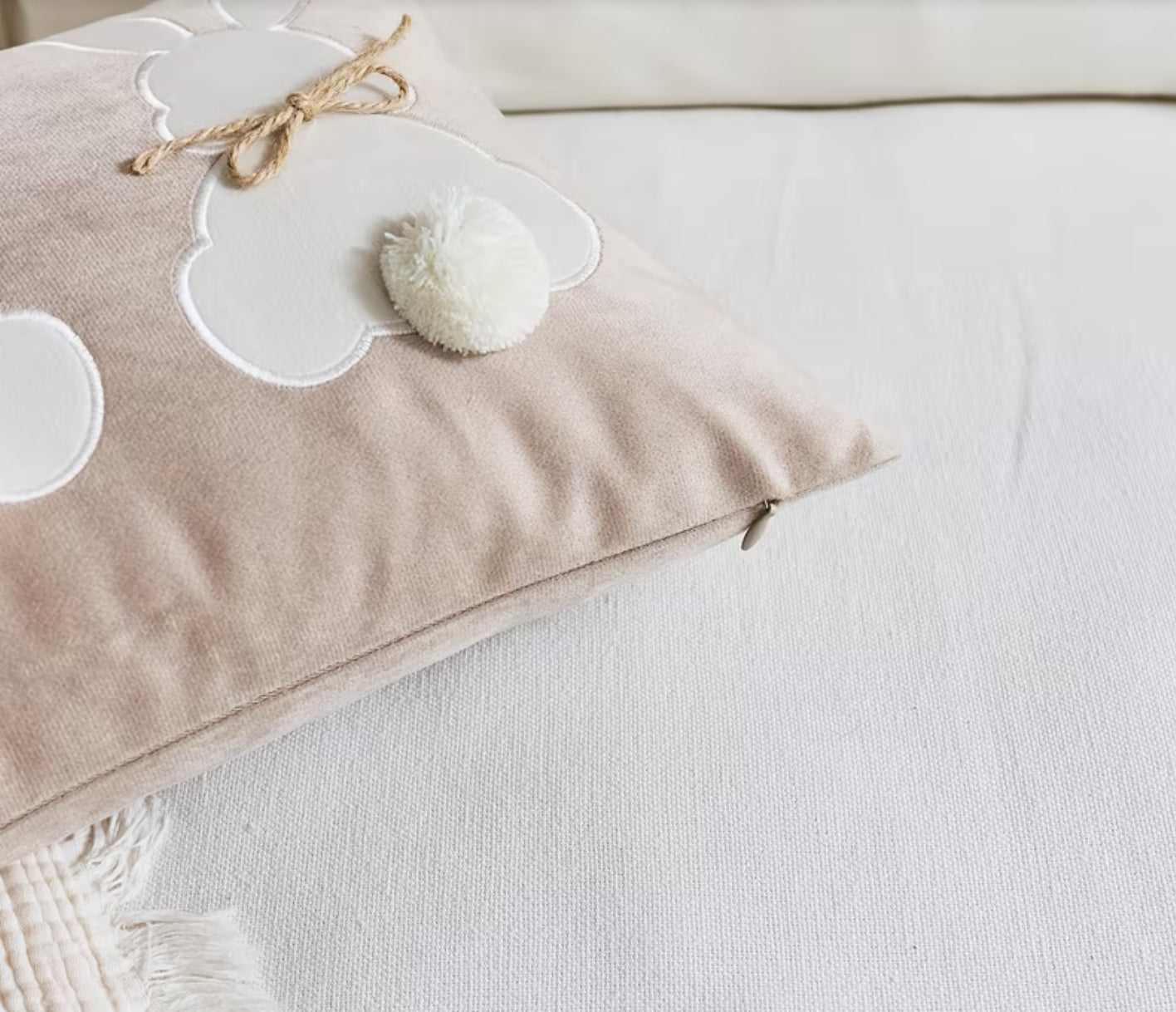 Beige pillow with white cloud designs and a pom-pom on a white surface