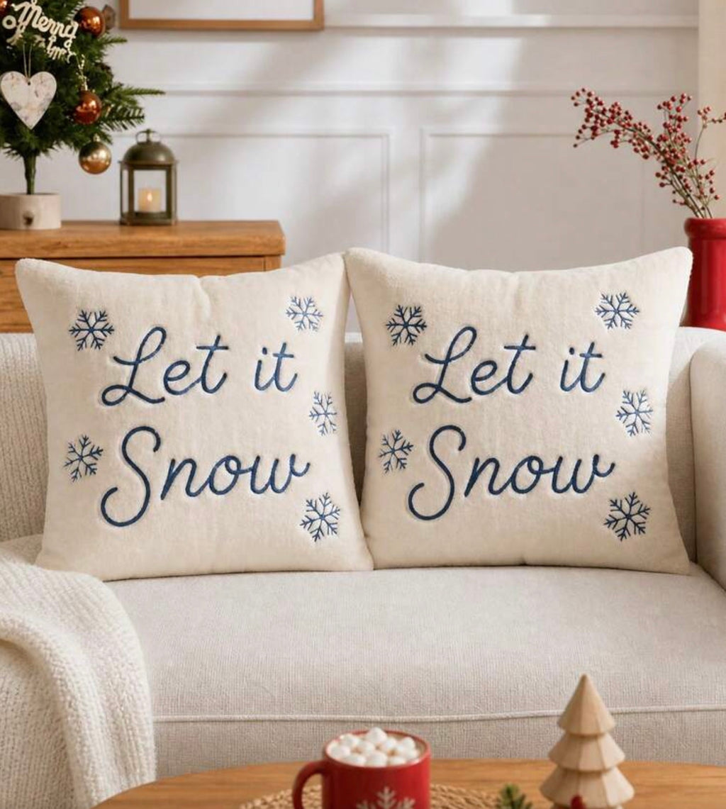 Two cream throw pillows with blue embroidered 'Let it Snow' text and snowflakes, displayed on a light beige sofa in a cozy living room setting.