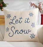 A square cream throw pillow with the phrase 'Let it Snow' embroidered in navy blue script, surrounded by four matching snowflake motifs. The pillow is styled on a light-colored sofa with holiday decorations in the background.