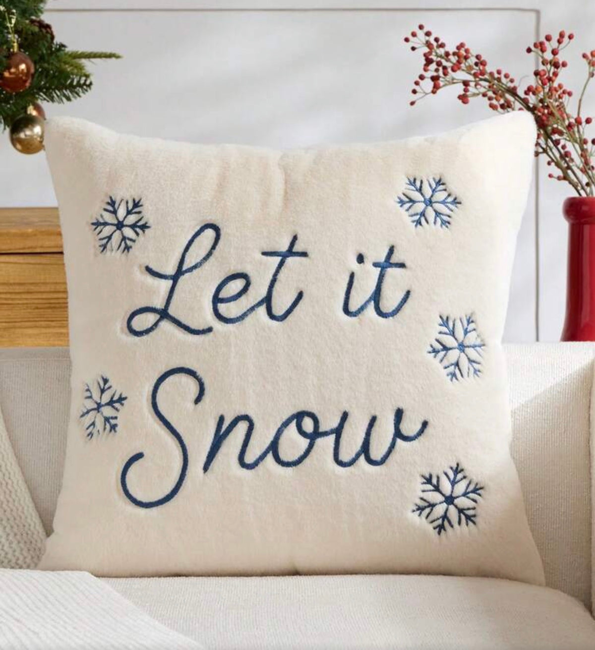 A square cream throw pillow with the phrase 'Let it Snow' embroidered in navy blue script, surrounded by four matching snowflake motifs. The pillow is styled on a light-colored sofa with holiday decorations in the background.