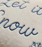 Close-up of a cream-colored plush throw blanket featuring embroidered navy blue script text that reads 'Let it snow' and a small snowflake motif in the corner.