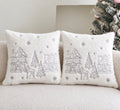 Two square white faux-fur throw pillows with embroidered winter village scenes, displayed on a beige sofa next to a decorated Christmas tree.