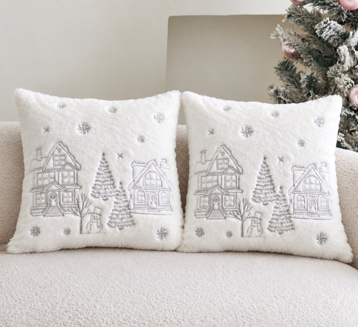 Two square white faux-fur throw pillows with embroidered winter village scenes, displayed on a beige sofa next to a decorated Christmas tree.