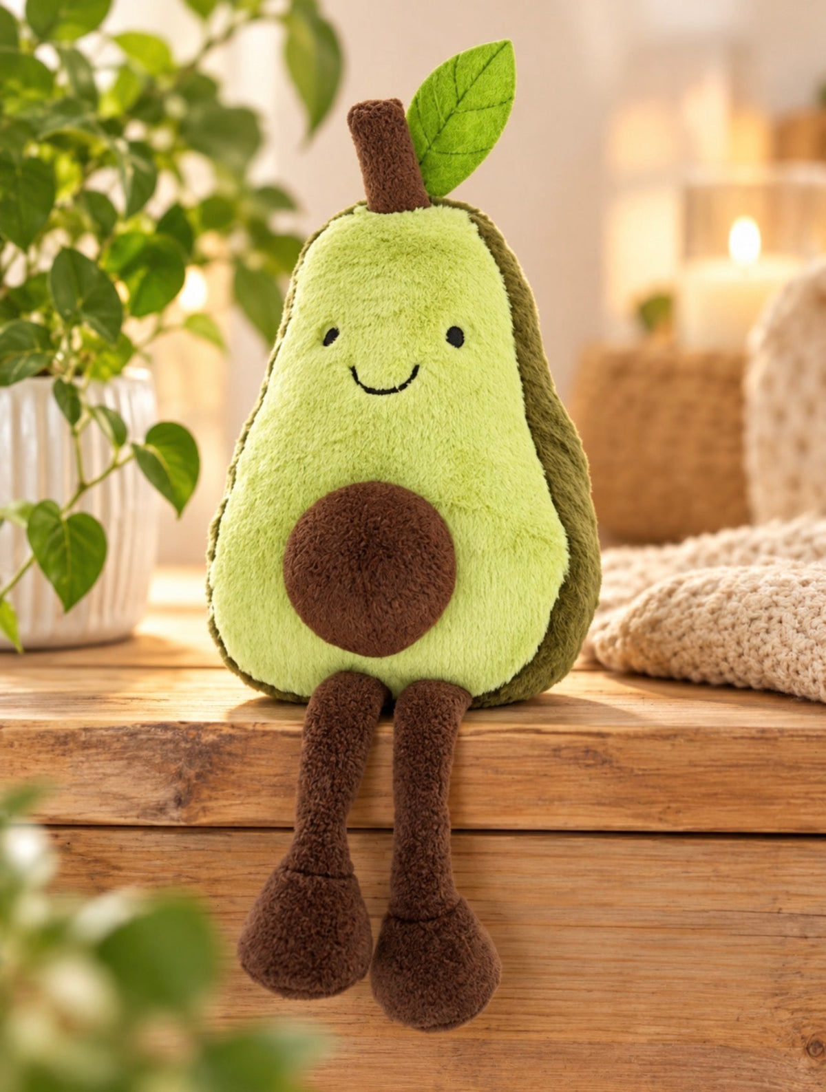 A plush avocado toy with a smiling face, green body, brown stem, and hanging legs, sitting on a wooden surface in a cozy indoor setting with plants and warm lighting in the background.