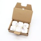Open cardboard box containing six white bath bombs with purple specks on a white background